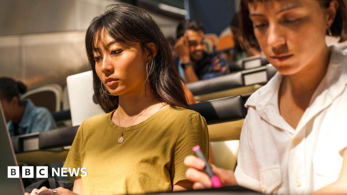Students sit in a bright lecture hall with tiered seating. Two female students are in focus. The one of the left wears a mustard-yellow T-shirt, a gold necklace, has long dark hair with a fringe, and large hoop earrings. She is typing on a laptop. The...