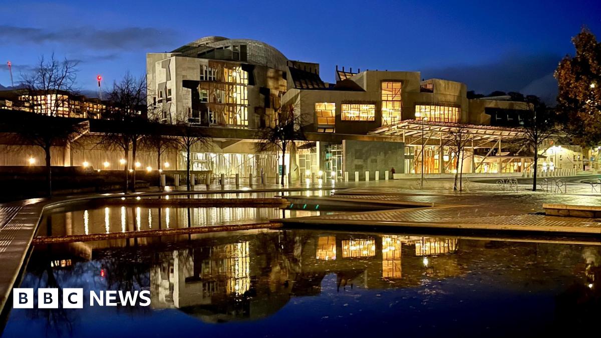 A scenic photograph of the Scottish Parliament building in Edinburgh, reflected in the ponds outside its entrance, taken at night by the author of the article. The illuminated building glows yellow against the dark blue of the evening sky.