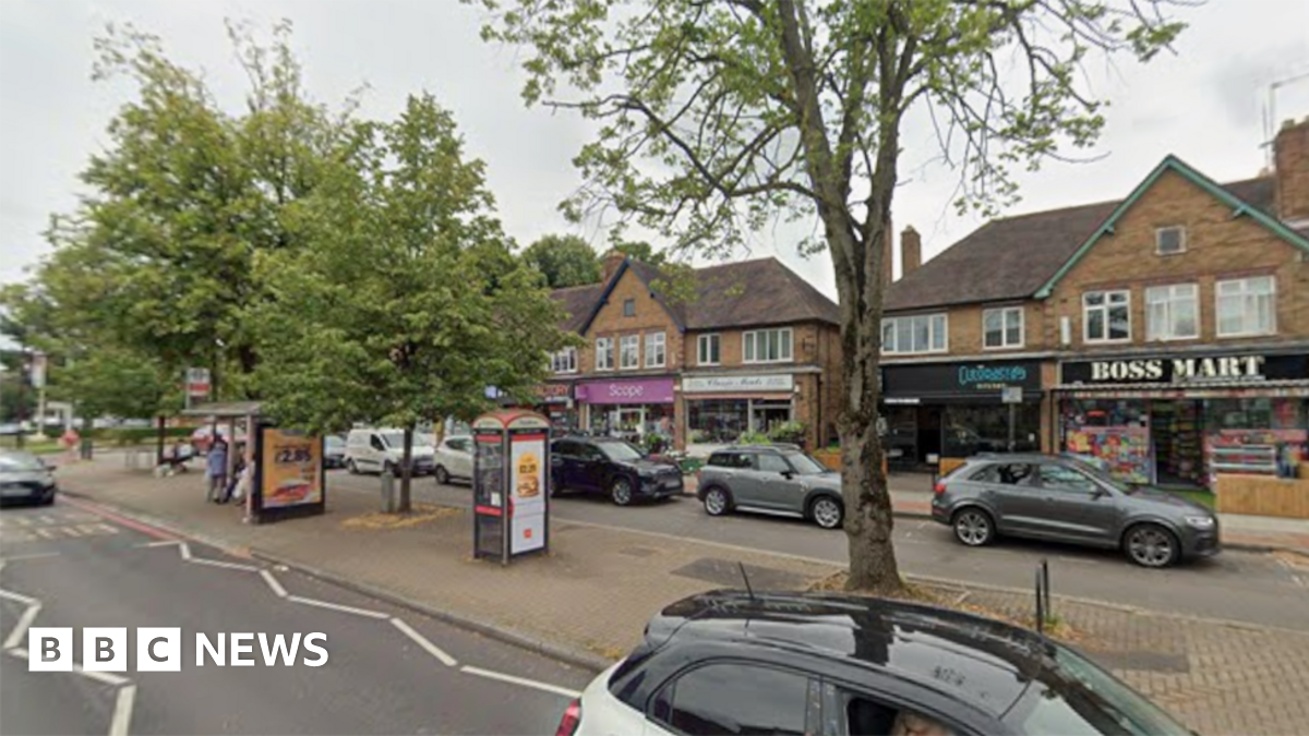 General view of a row of shops on Stratford Road, Shirley. In the background is a row of shops, in front of which are parked cars. A central reservation has a bus shelter, phonebox and trees, with other traffic passing in the foreground.