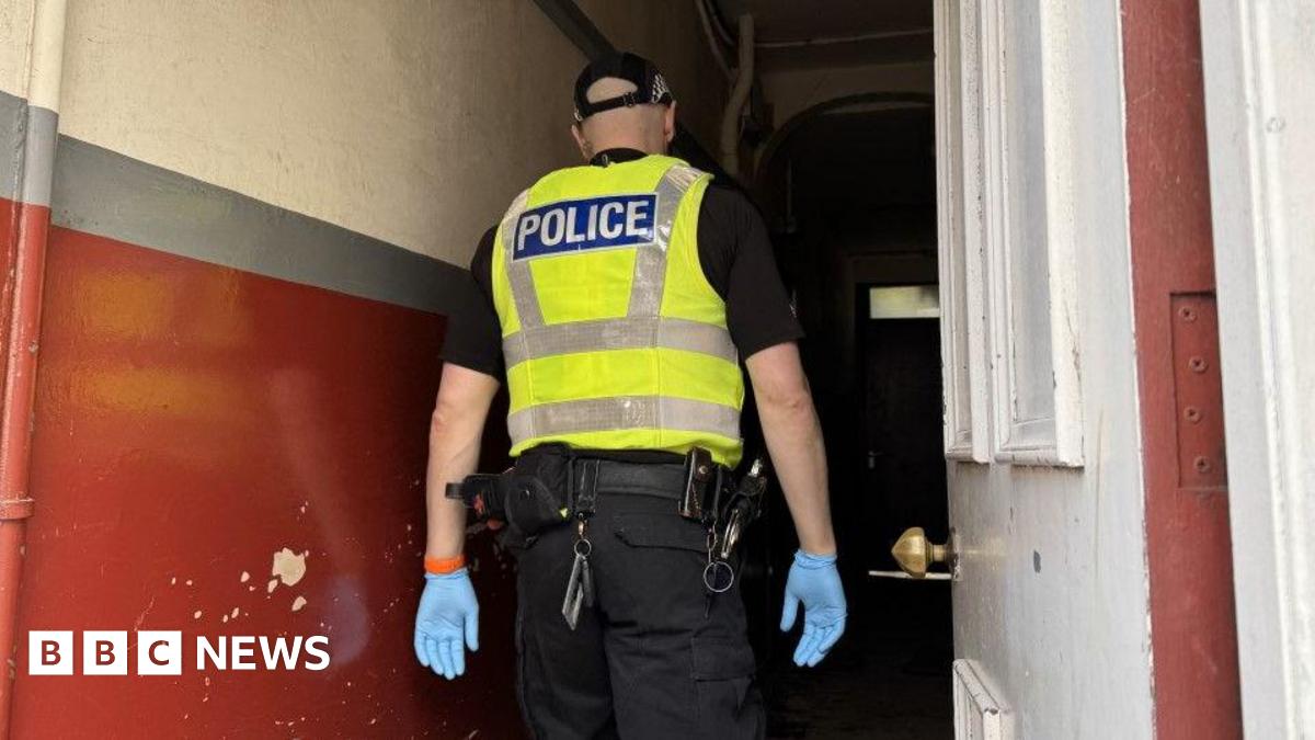 A Police Scotland officer in black uniform with a yellow hi-vis tabard and blue latex gloves walks into a painted close. The walls are burgundy and cream. A white main door is open to the right.