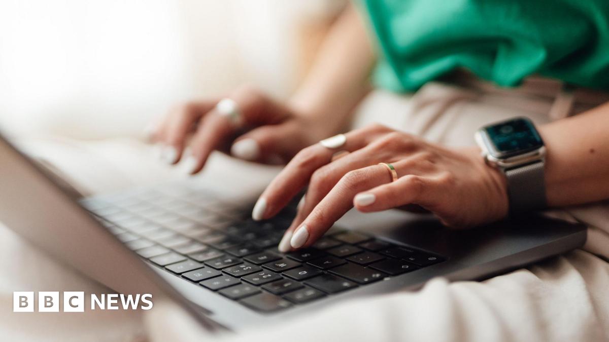 Someone's hands typing on a laptop keyboard. they have white nails and are wearing gold jewellery. They have an apple watch on their rest and are wearing a green top and beige trousers. The laptop is silver.