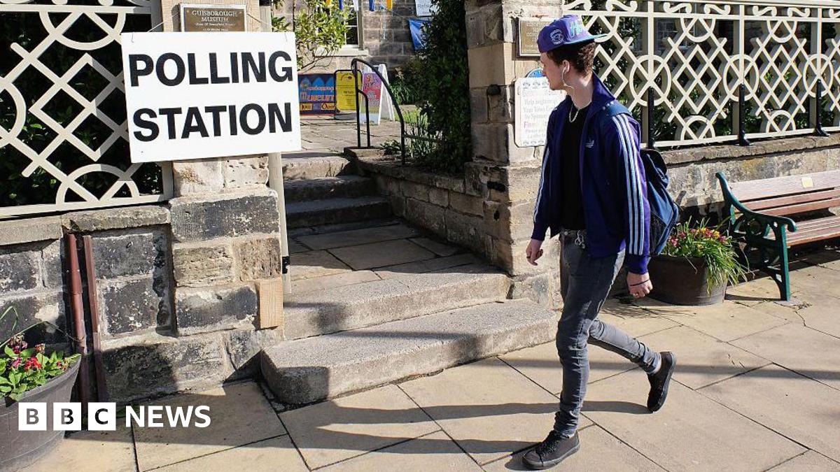 A young man wearing a cap and listening to headphones walks past a sign reading "polling station".