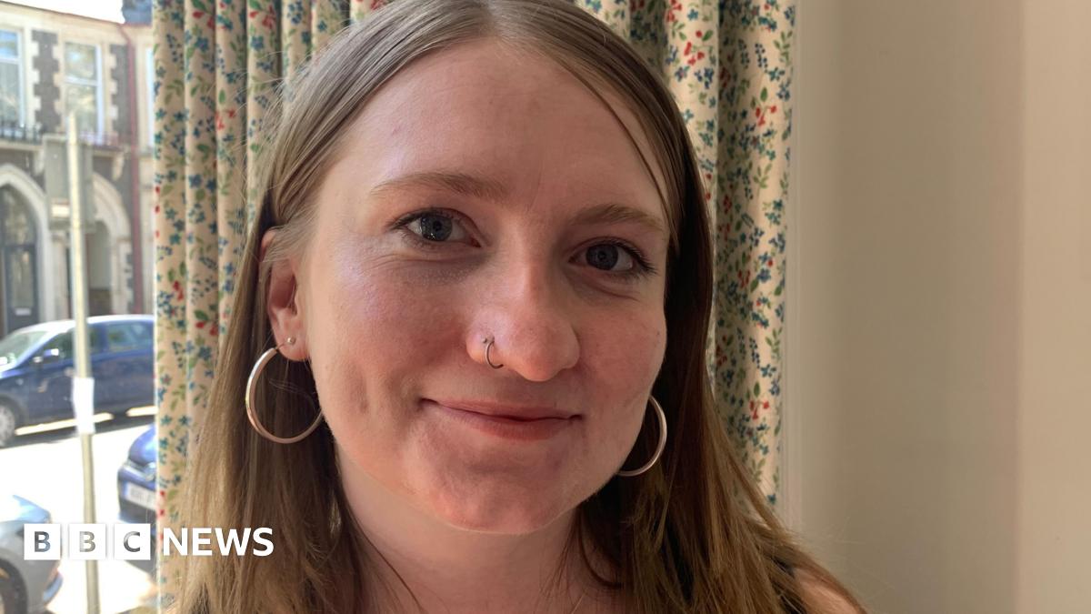 Marianne looks at the camera, she's wearing hoop earrings and has a nose piercing. She's smiling and a curtain and window overlooking the street is visible behind her.