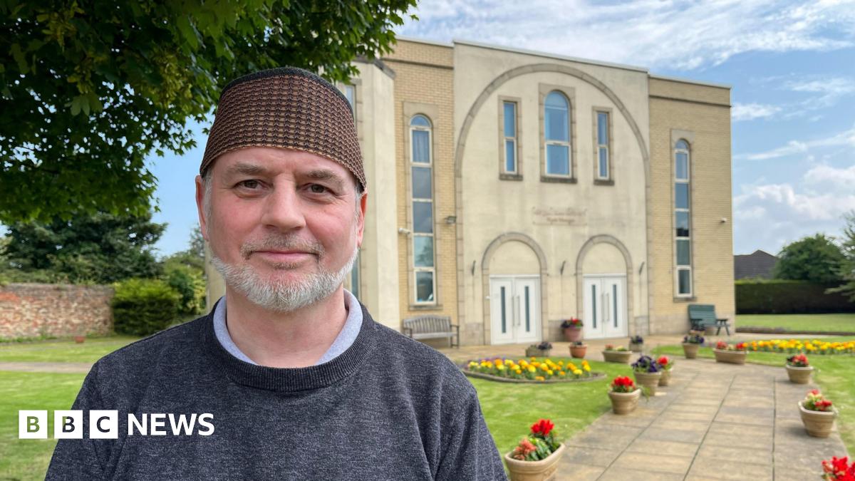 A man, smiling, in front of a mosque; he has a short, white beard and is wearing a taqiyah cap.