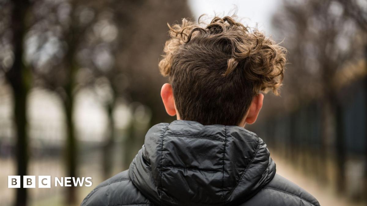a teenager, with short curly dark hear and wearing a black jacket is facing away from the camera
