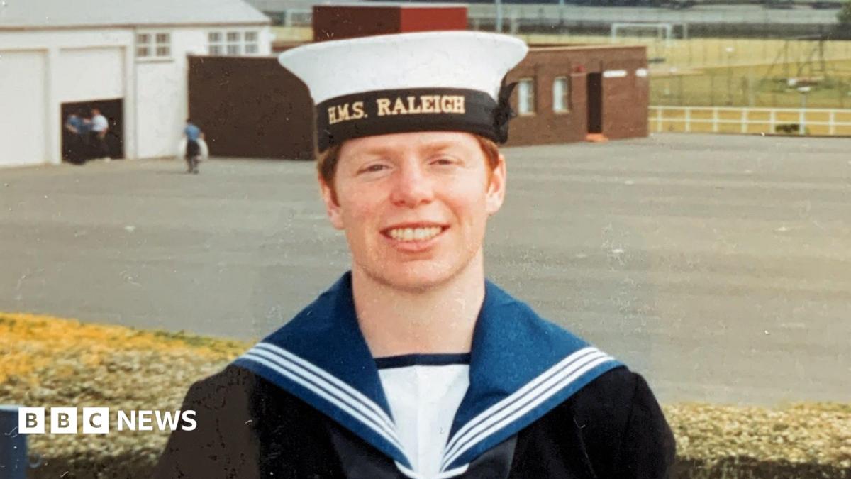 Jennifer stands in her uniform as a young woman. She appears to be on base, and wears a white hat with a black rim, and black jacket with a white shirt and a blue collar with white stripes on it. She has red hair and is smiling directly at the camera. 