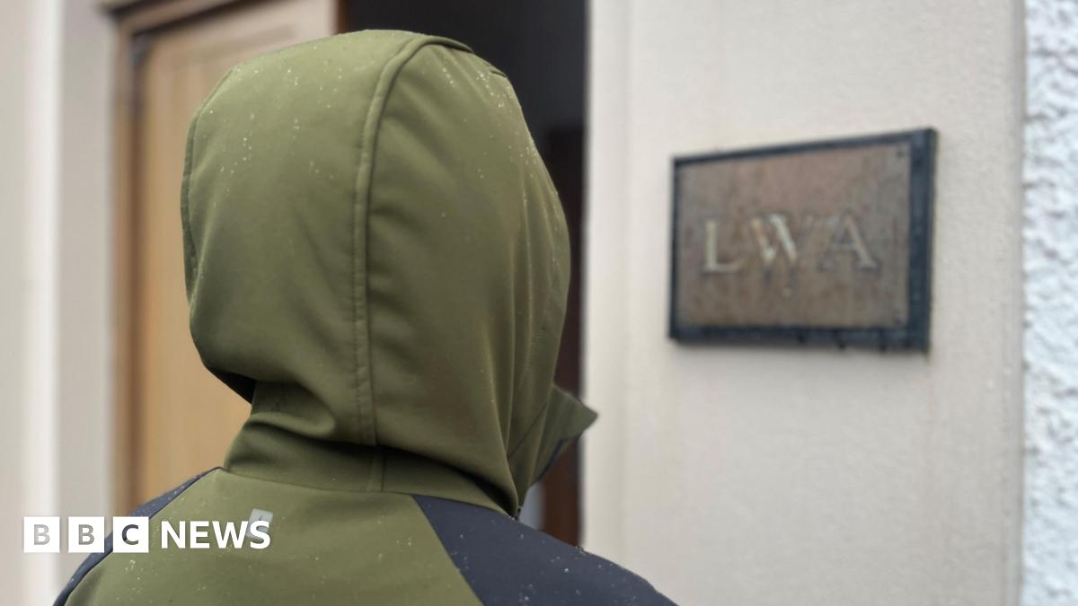 Unidentifiable person with their back to the camera wearing a khaki waterproof jacket with the hood up. They are standing outside a building with a sign that says "LWA".