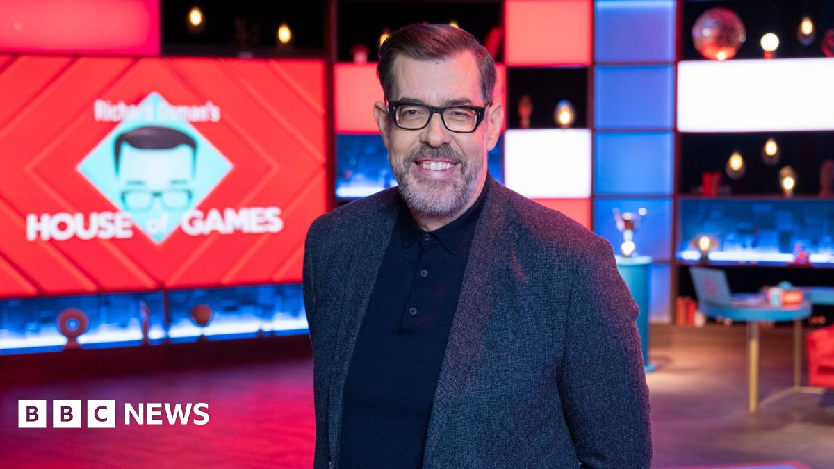 Richard Osman, wearing a dark suit and black glasses, on the House of Games red and blue set