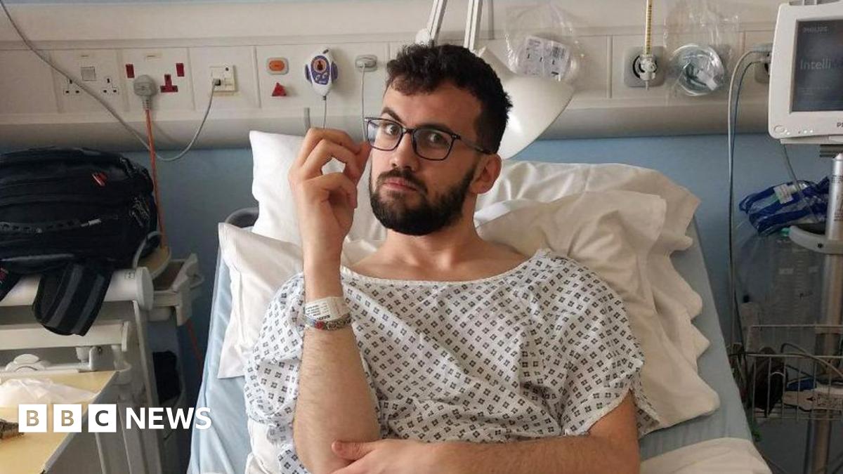 Chris Brookes-Smith in a hospital gown in a hospital bed. He has a beard and glasses