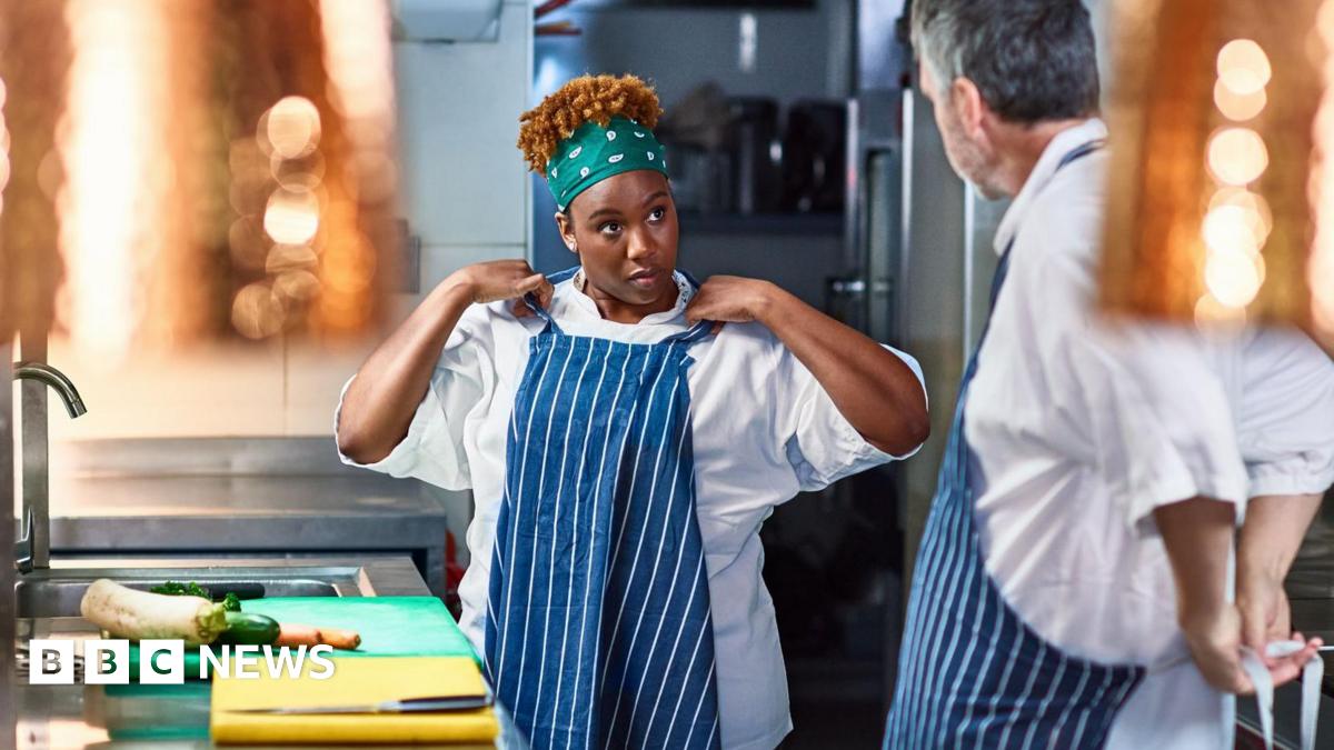A stock photo of a trainee chef adjusting her apron in a kitchen while looking at her older mentor. There are ingredients on the counter in front of her.