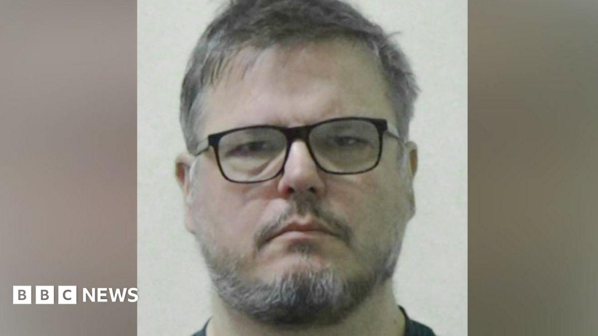 Mugshot of Dean Juric. He has scruffy dark and grey hair and a black and grey beard and is wearing black-framed glasses