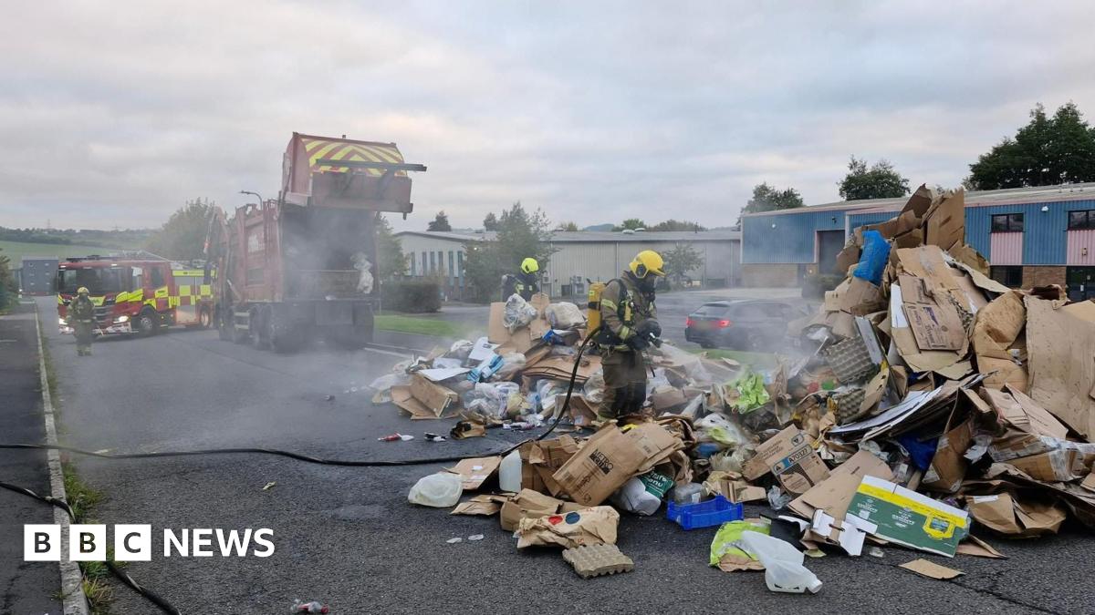 Two firefighters pour water from hoses on a large pile of cardboard and other recycled material while dealing with a fire. Smoke is coming from the pile. The lorry the items were in is parked near the firefighters. A fire engine is next to the lorry.