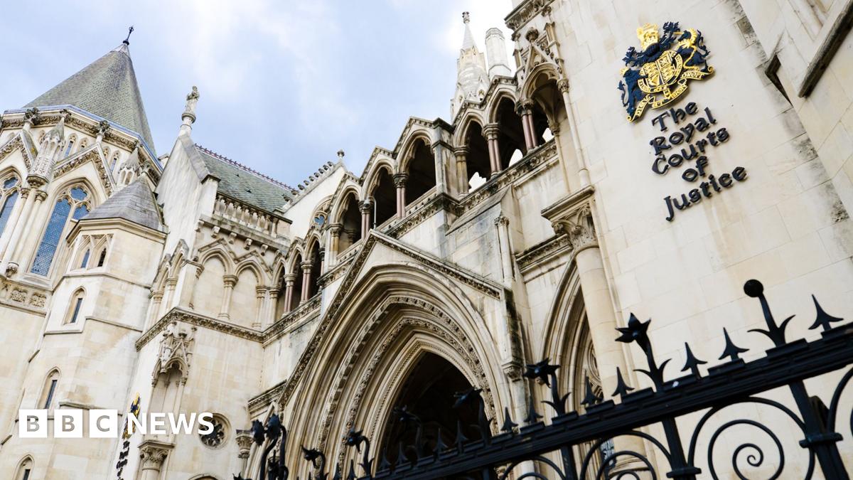 A general view shows a side-on view of the Royal Courts of Justice in London, with a sign on the side of the building showing the coat of arms and a blue cloudy sky in the background.