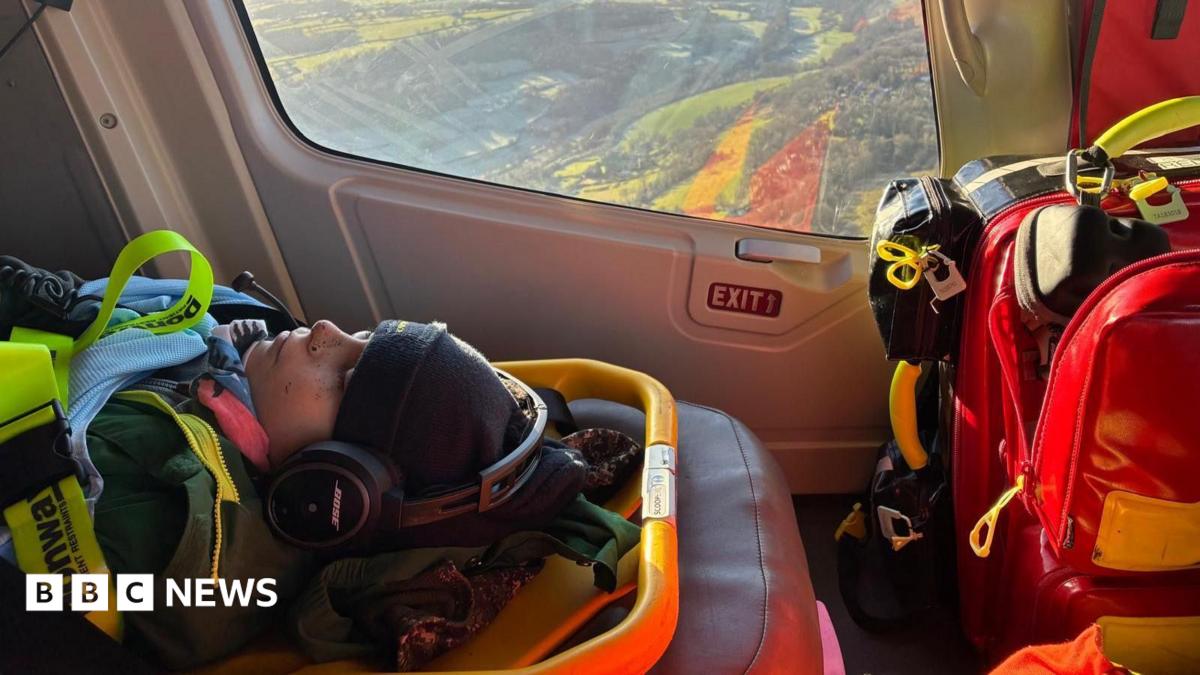 Inside a helicopter, the window in the background showing the landscape below. In the foreground, Dom, 12, lies unconscious on a stretcher with hat and coat and lots of straps to keep him in. There is a large bag of equipment to the right.