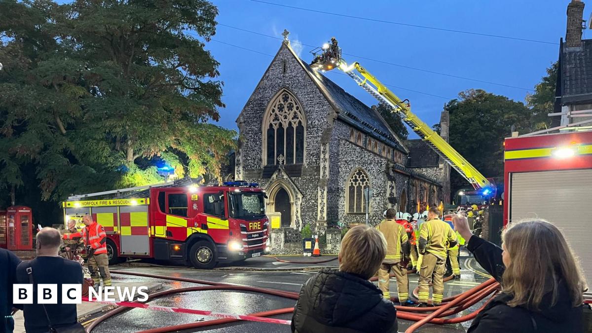 Firefighters on a ladder just above the roof of a church, just behind its pointed gable end. A small amount of smoke is behind the tip of the gable end, which has a cross at its apex. Fire engines and passers by are in the street. There are red hoses a...
