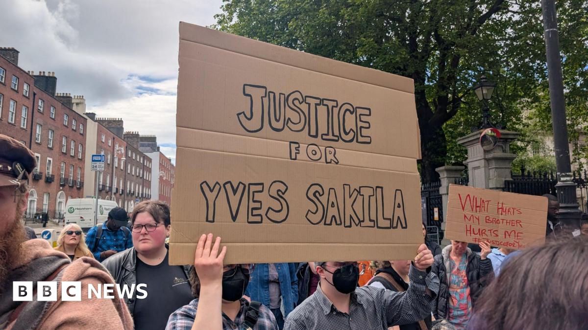 Two people wearing black face masks, hold up a cardboard sign which reads 'Justice for Yves Sakila' in black writing. They stand among a group of people in the shade of large green trees.