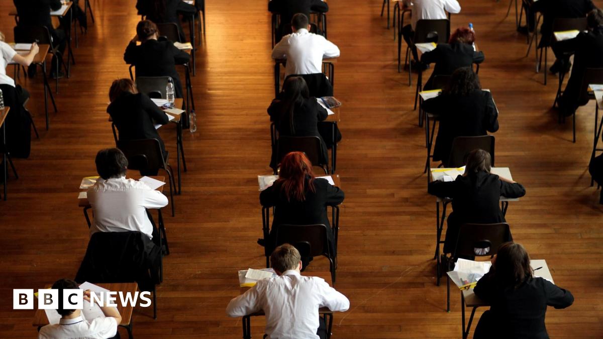 View from behind of pupils doing exams in a school hall. They are sitting at separate desks which have been spaced out for exam conditions. There are papers on their desks. Some of them have got black jumpers on while others are just in white shirts.