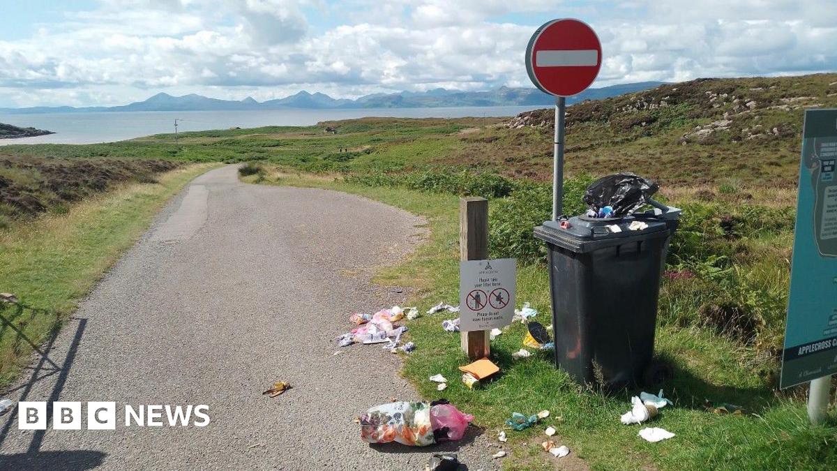A wheelie bin overflowing with litter, next to a "No Entry" sign. It sits next to single track road which leads off into the distance. In the background is a stretch of water and a picturesque, mountainous landscape. 