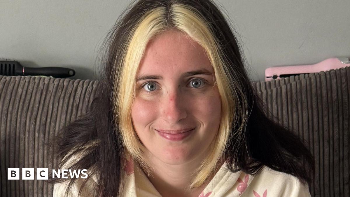 A close-up photo of Kailee who looks into the camera with a smile, she is sat on her sofa at home. She has dark hair down over her shoulders, apart from the front which is dyed blonde.