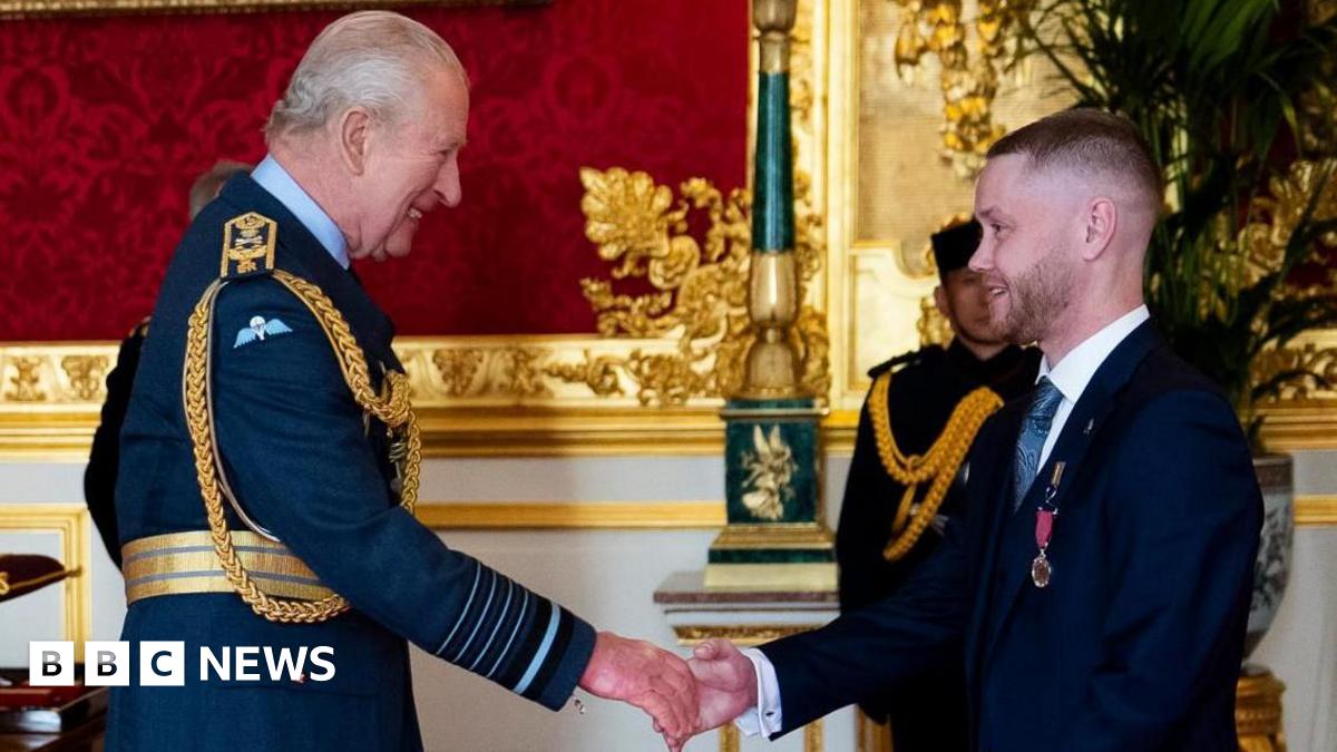 King Charles in RAF uniform smiles at Nathan Newby, a man in a dark blue suit, as he pins a medal on him. In the background is another man in military uniform who stands in a grand-looking room with gold decorations and a carved marble fireplace.
