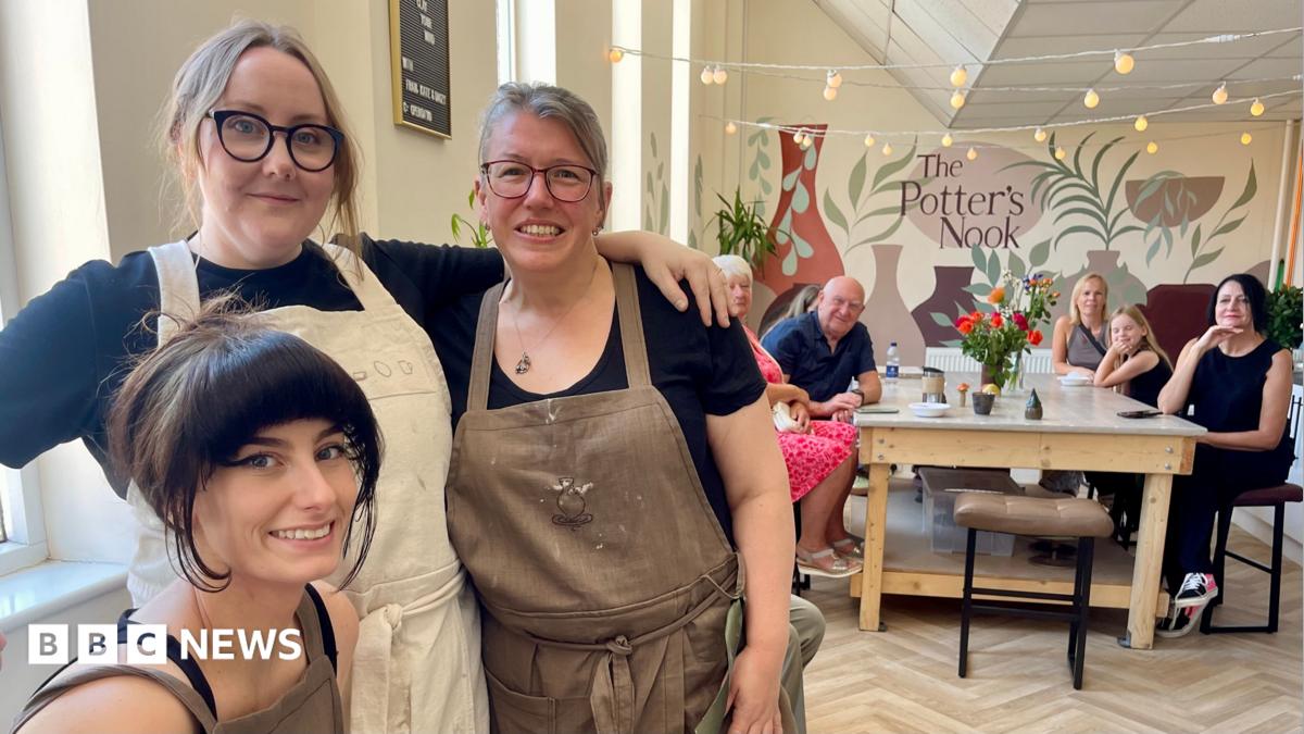 Three women in black tops and brown aprons are in a pottery studio. There is a large table which four people and one child are sat at. There is a large mural on the back wall which includes a bottle kiln and the words “The Potter’s Nook”