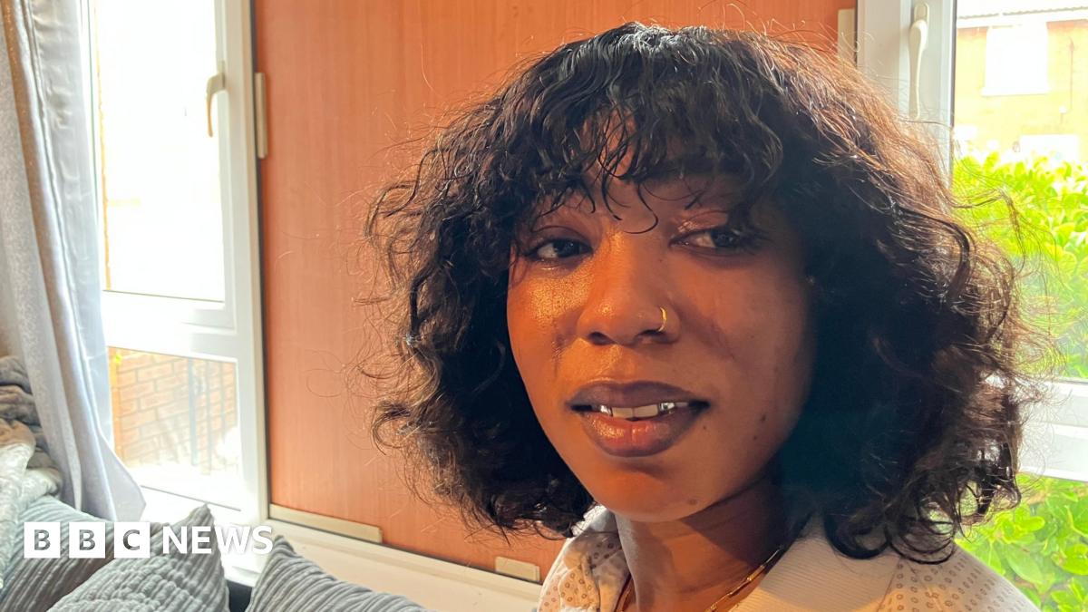 Sia Fay has shoulder-length curly hair and a nose ring. She is wearing a cream polo shirt and is sitting on a light grey/blue sofa. The windows of her front room are behind her, one is boarded up.