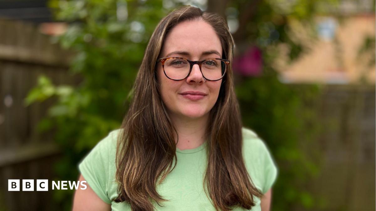 Erin wearing a green top and glasses. She is looking direct at the camera and standing in a garden. She has long brown hair hanging to chest level.