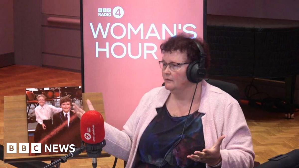 A woman with brown curly hair wearing a dark blue dress and pink cardigan, during an interview for BBC Woman's Hour. She wears headphones and is in front of a microphone with the Woman's Hour logo behind her. There is a photo of two boys next to her.