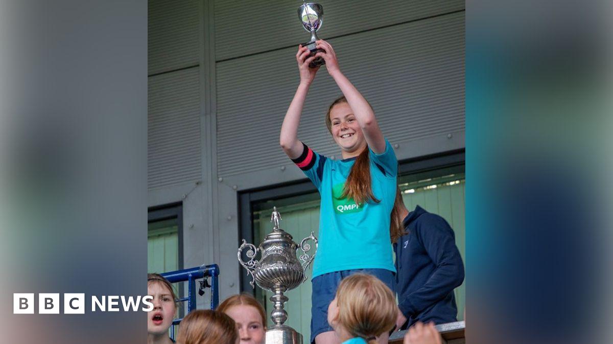 Clara has long brown hair in a ponytail. She is wearing a light blue football shirt. There is a large trophy beside her. There are children in the foreground.