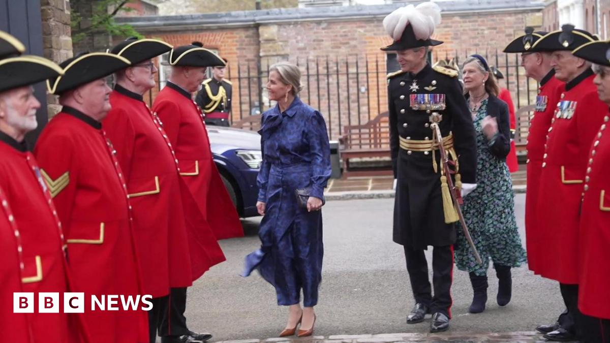 Chelsea Pensioners in their distinctive red tunics stand in two rows as an honour guard as the Duchess of Edinburgh walks into the Royal Hospital, accompanied by a man in formal uniform with rows of medals on his chest. He is wearing a plumed hat. 