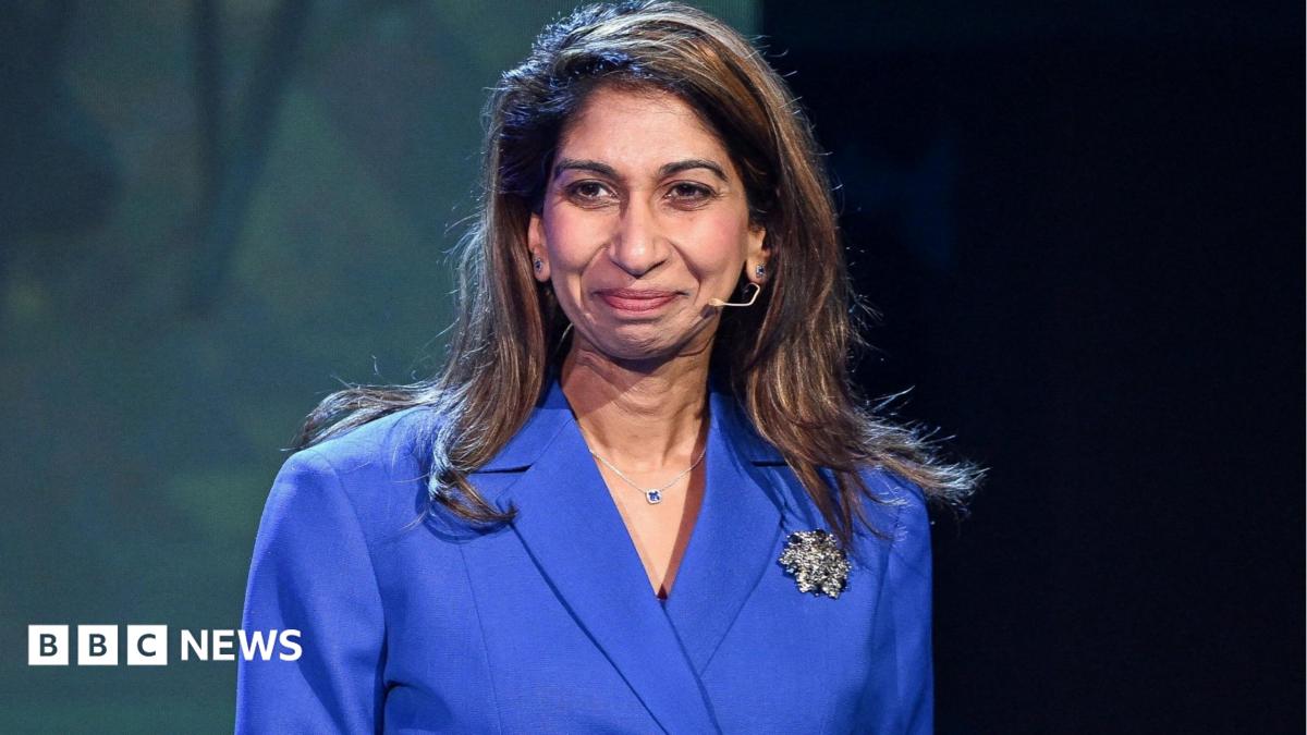 Suella Braverman is standing in front of a backdrop and in front of a black podium. She has brown hair and is wearing a blue suit jacket with black buttons. She has a large silver brooch on the jacket and a nude coloured microphone wire near her mouth
