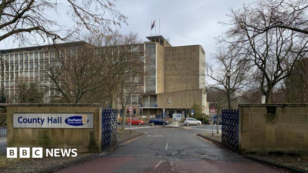 An outside view of Durham County Hall from outside the gates. Cars can be seen parked outside the large grey building.