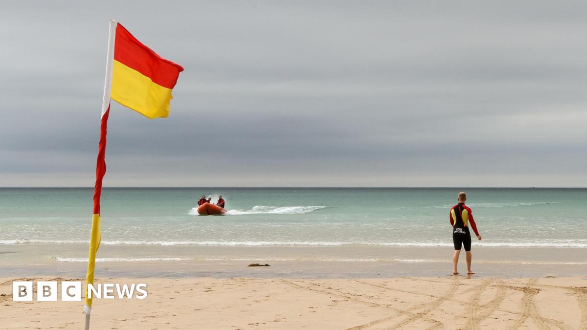 A beach showing a flag marking the safe swimming area. A lifeguard is stood on the beach while looking at a rescue boat heading towards shore. 