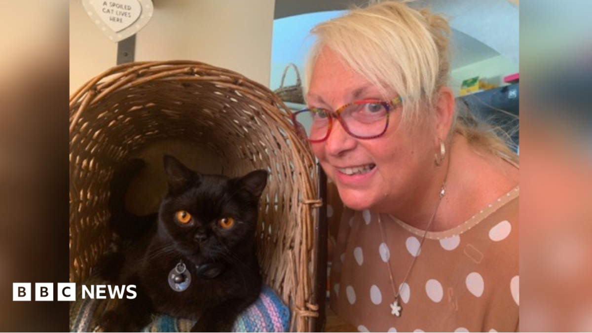 The image shows a woman smiling while posing with her black cat, who has amber eyes and is sitting in a basket.