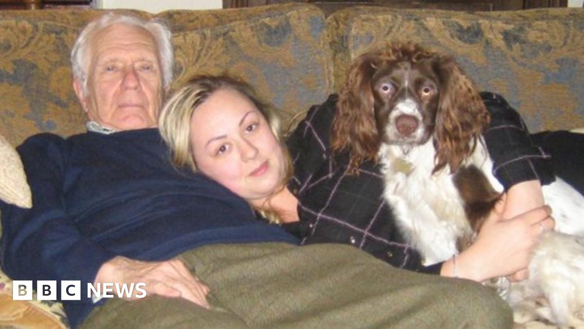 John Wilcox, an elderly man, is leaning back on a sofa, wearing a dark blue pullover and green trousers. His granddaughter Amy is lying across the sofa, her head on John's stomach. She is holding a brown and white dog.