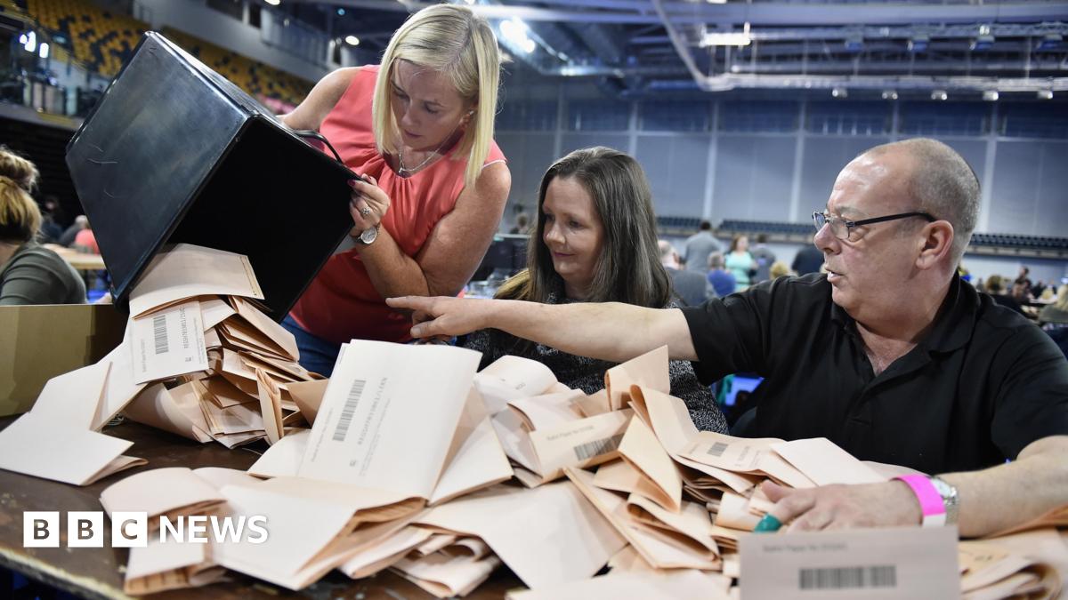 Counting staff check ballot papers in the Scottish Parliament elections at the Emirates Arena, Glasgow, in 2016. A woman on the left is pouring a box of pink papers onto a table, which is covered with ballot papers. A man on the right is pointing at th...