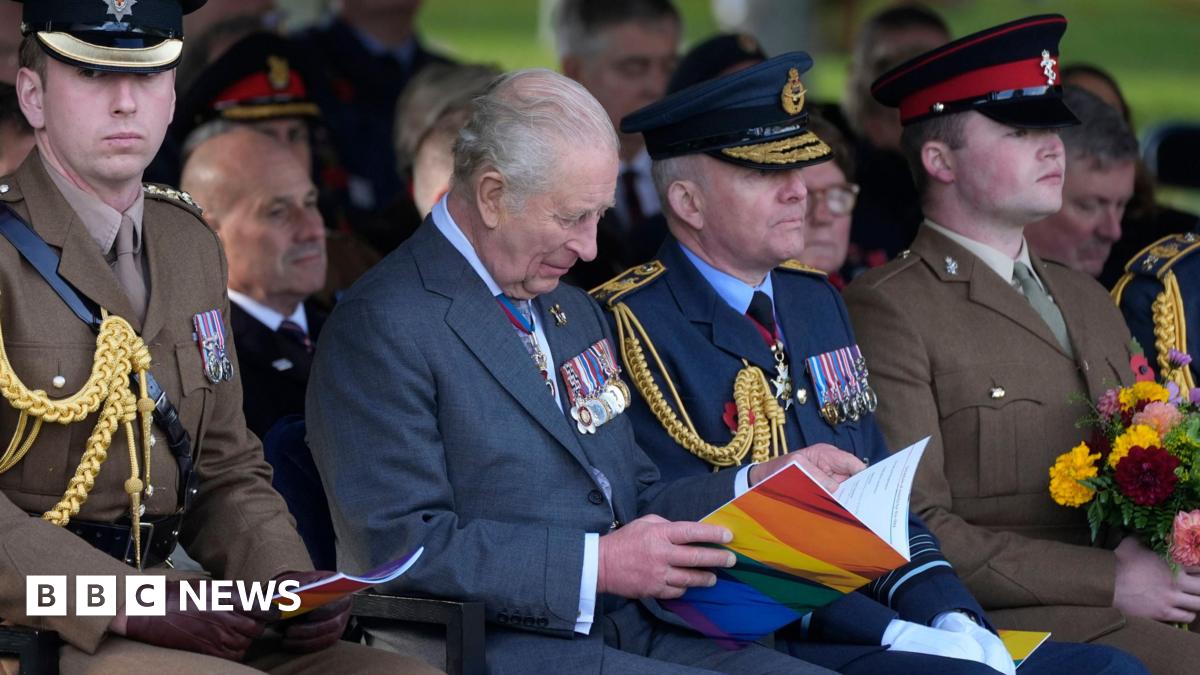 The king sits in the front row of the dedication ceremony flanked by uniformed members of the armed forces. He is looking down reading an open book of service with the LGBT rainbow on the front.