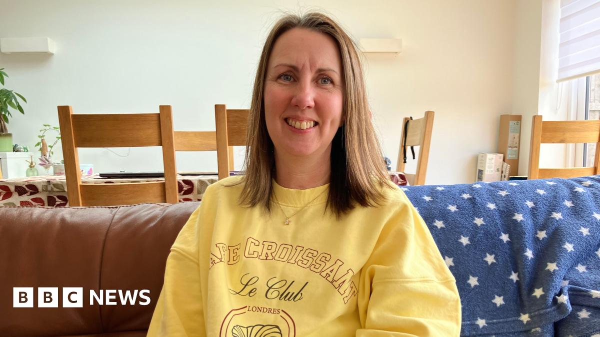 A woman with medium-length brown hair wearing a bright yellow jumper smiling, she is sat down on a brown leather sofa with a blue and white stars blanket draped over it. 