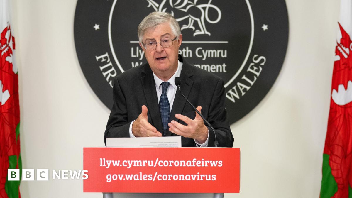 Mark Drakeford, delivering Covid updates to the public during the pandemic. He is standing at a podium, in front of the Welsh government sign, with a Welsh flag on either side of him.