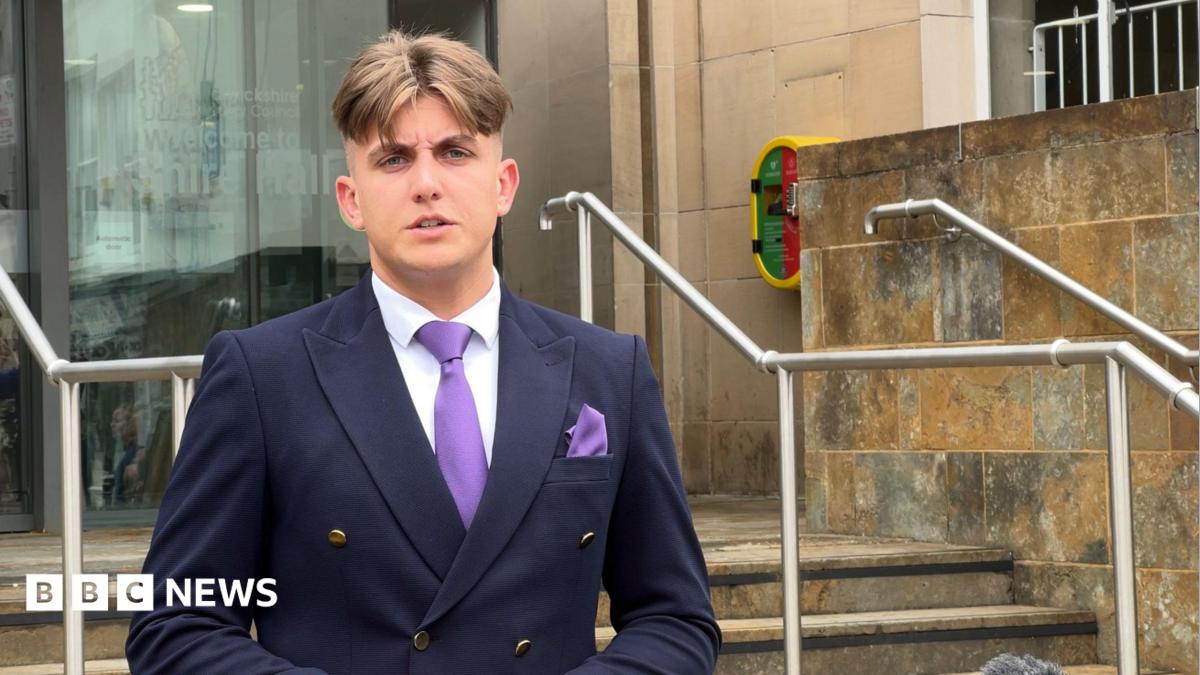 A man standing in a navy blazer and purple tie, on the steps of a council building built in light sandstone.