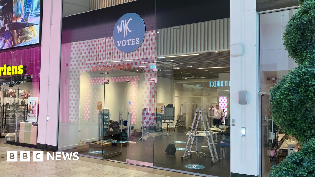 A glass-fronted polling station inside a shopping centre, marked with a circular “MK Votes” sign, with voting booths and a ladder visible inside.