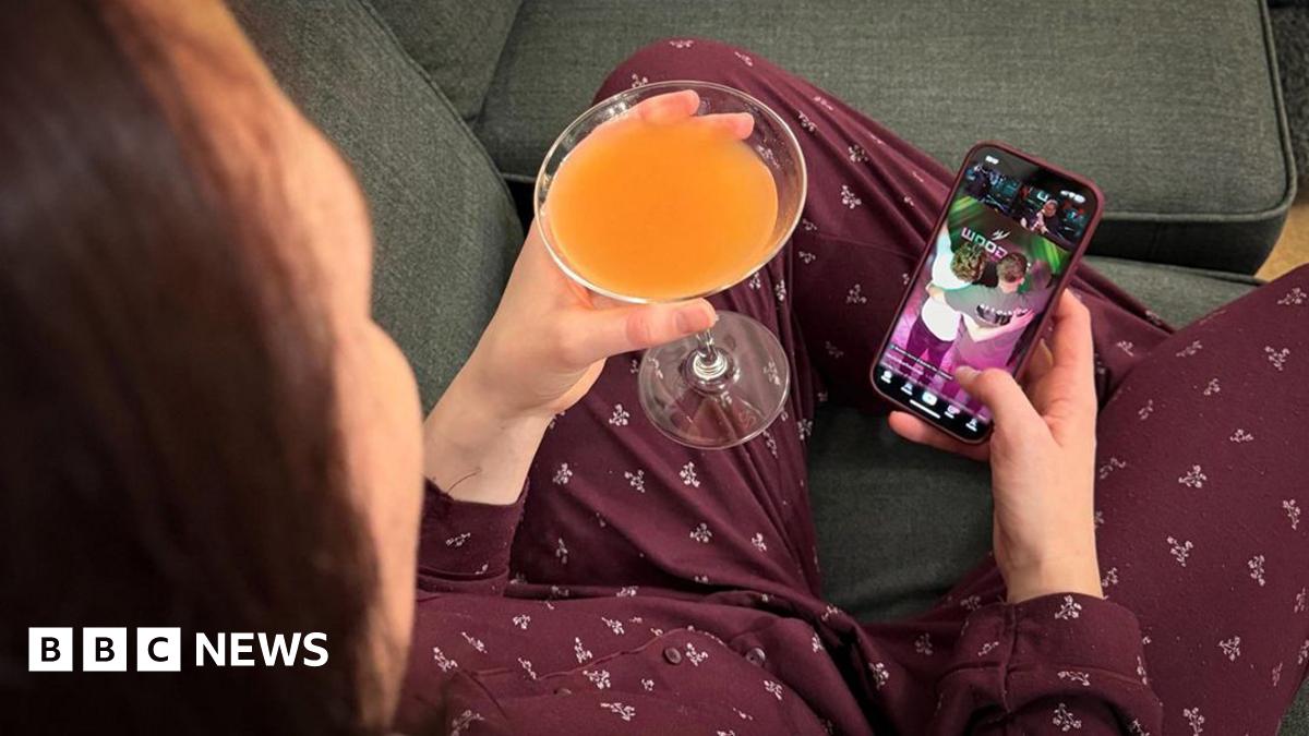 Woman sat on a sofa holding a cocktail in one hand and her phone in the other. She is watching a live stream from a bar in Manchester.  (Stream image has been added to phone by Photoshop for illustration purposes)