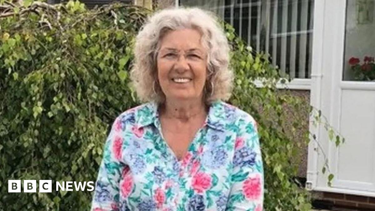 Valerie Kneale with shoulder-length grey curly hair wearing a pink, lilac and green floral blouse smiling in front of a shrub. She is standing in front of a pebble-dashed house with a white plastic door and windows.