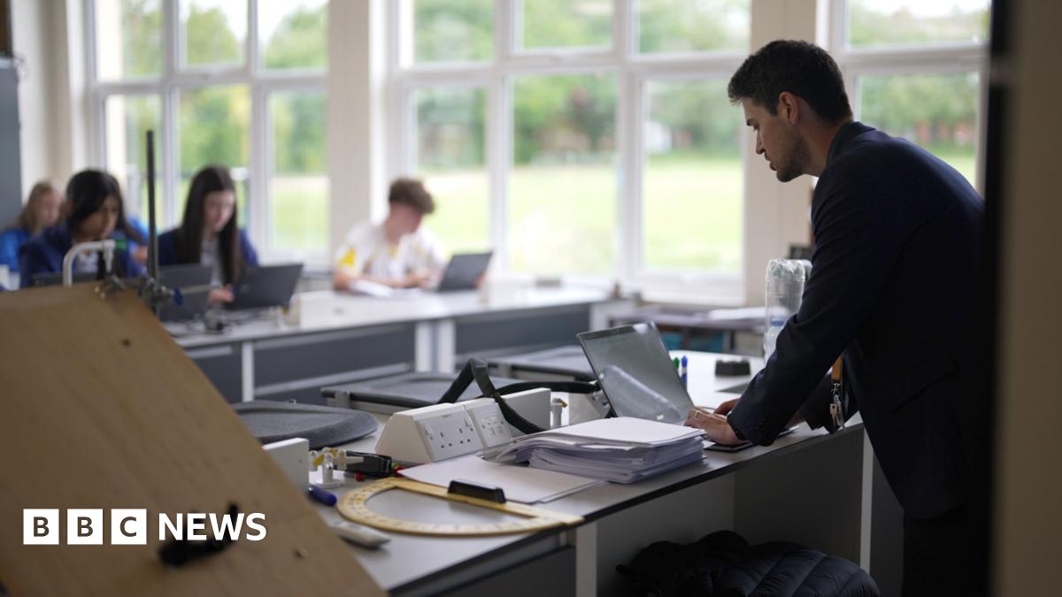 In a classroom of pupils, the male teacher in the foreground is looking down at his desk. In the blurred background are a number of students at their desks, with large windows opening out onto what appears to be a school field.
