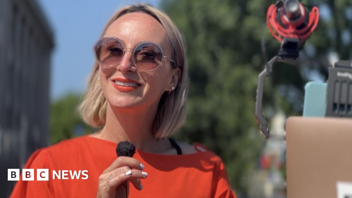 A woman with blonde hair in a red dress and sunglasses stands looking at the camera while holding a microphone.