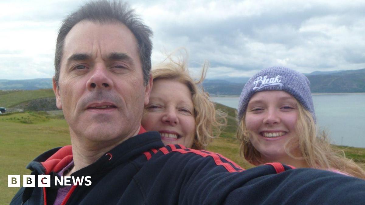 A picture of a man, woman and a teenage girl smiling at the camera standing on top of a hill. Mountains and the sea are in the background.