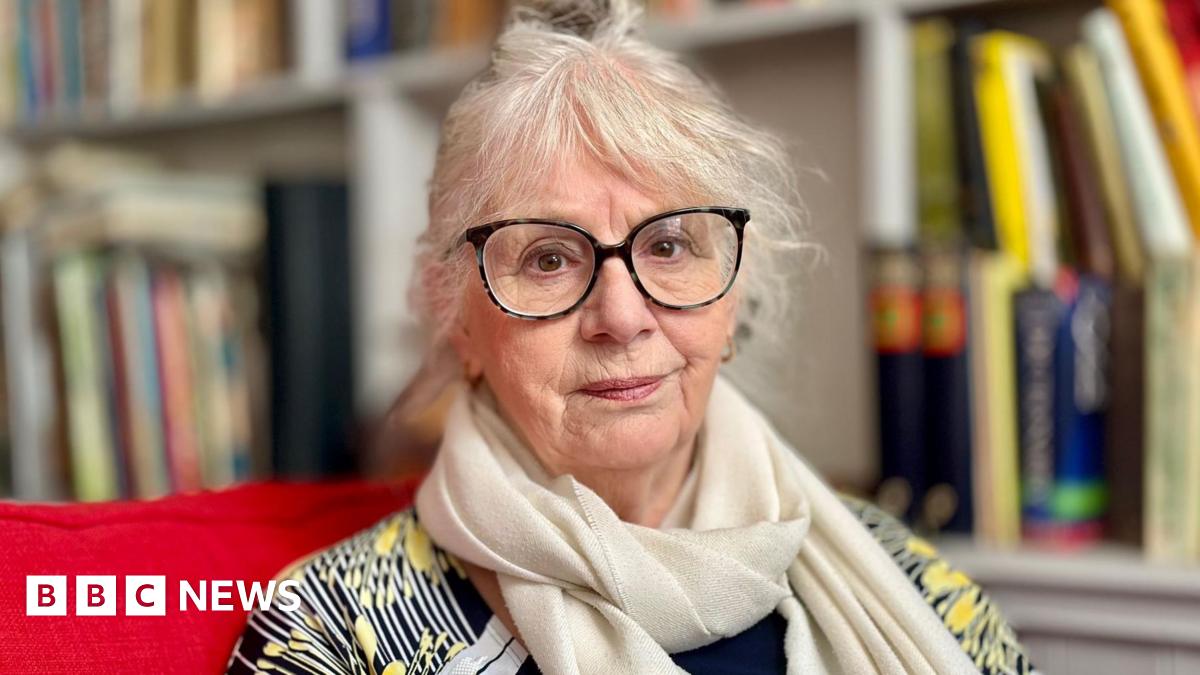 A woman aged 75 with white hair tied back, wih a fringe and large black coloured spectacles. She wears a cream scarf and a yellow and black patterned top. Bookshelves are visible behind her, she sits on a red sofa. 
