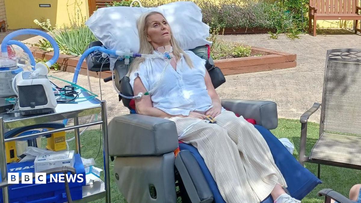 Catherine lying in a medical bed in a garden. She is connected to medical equipment. A young boy is sat by the bed.