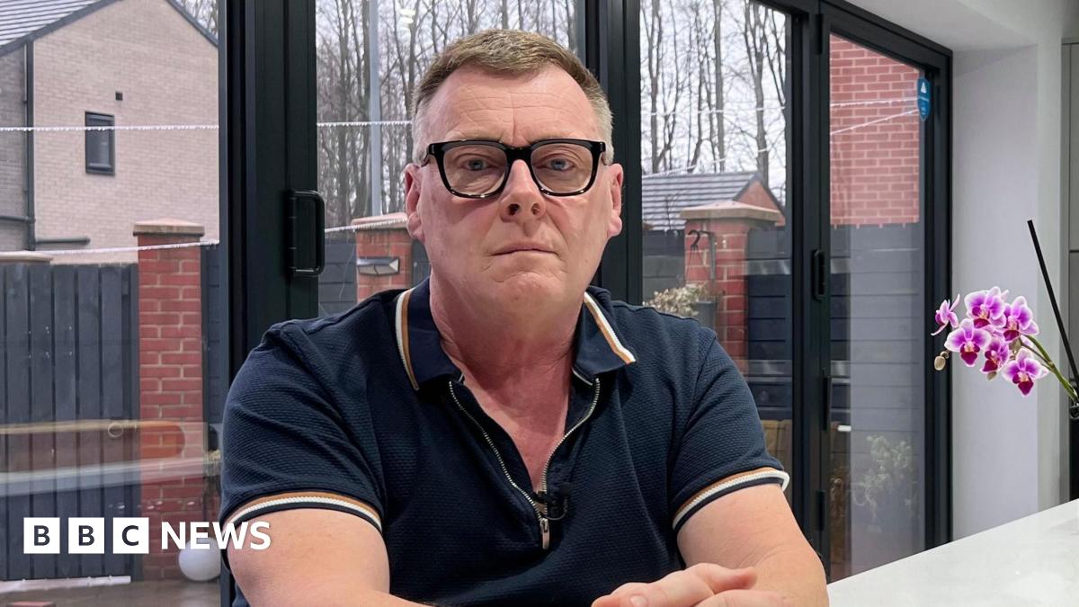 Graeme Blenkiron, a man with glasses and a navy polo shirt, is sitting at a table next to some cut flowers and an orchid.