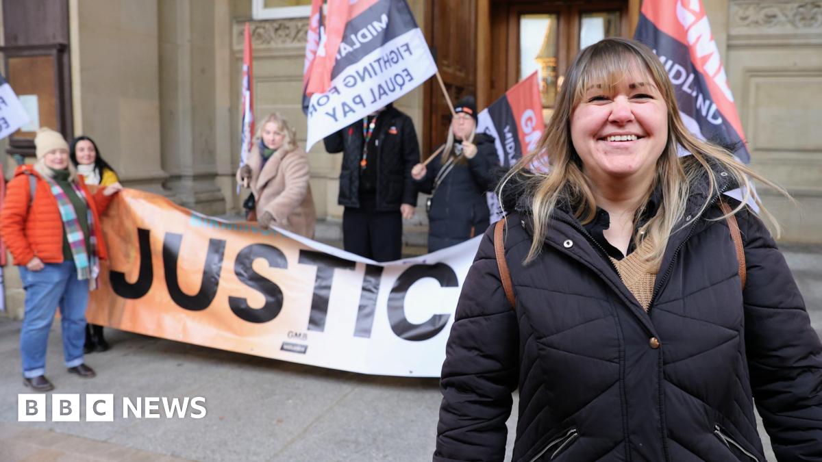 A woman in a black puffa coat standing outside a sandstone building with ornate carving on some bricks and stone pillars. Behind her stand four people holding a banner and flags saying "Justice and Fighting for Equal Pay"